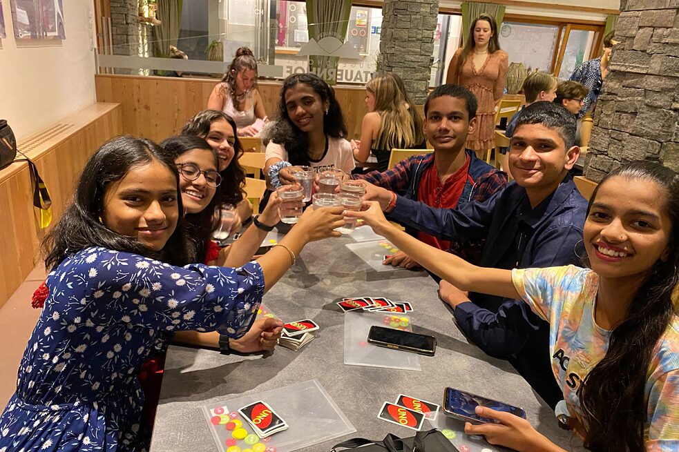 Jugendliche sitzen um einen Tisch herum, sie stoßen an. UNO Spielkarten liegen auf dem Tisch. 