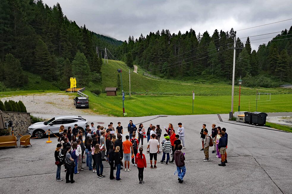 Eine Gruppe Jugendlicher steht auf einem gepflasterten Hof; im Hintergrund bewaldete Berge.