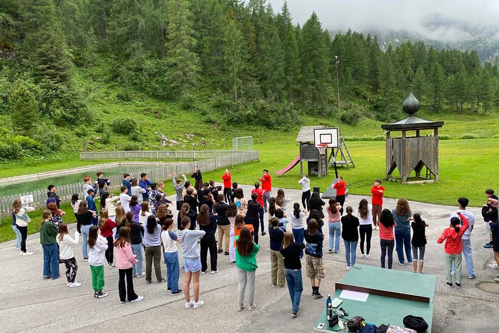 Jugendliche auf einem gepflasterten Hof bei Tanzbewegungen. Im Hintergrund sind bewaldete Berge zu sehen.