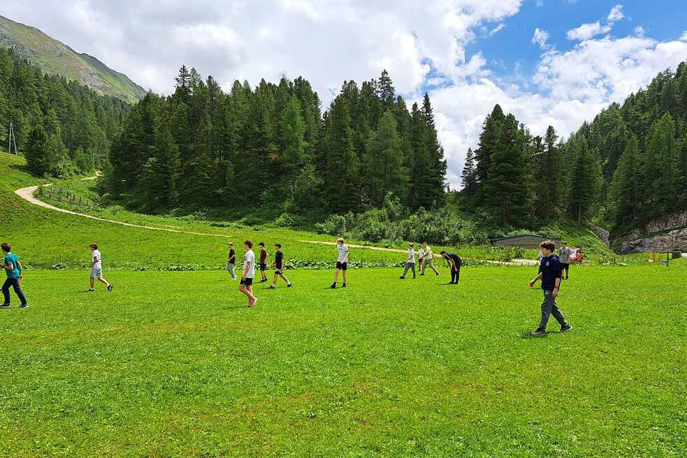 Zwei Jungs spielen Ball auf einer großen Wiese. Im Hintergrund Berge und Wälder.