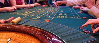 Roulettetisch mit einem langen, mit grünem Tuch (Tapis) bespannt, Jetons und Hände im Hintergrund.