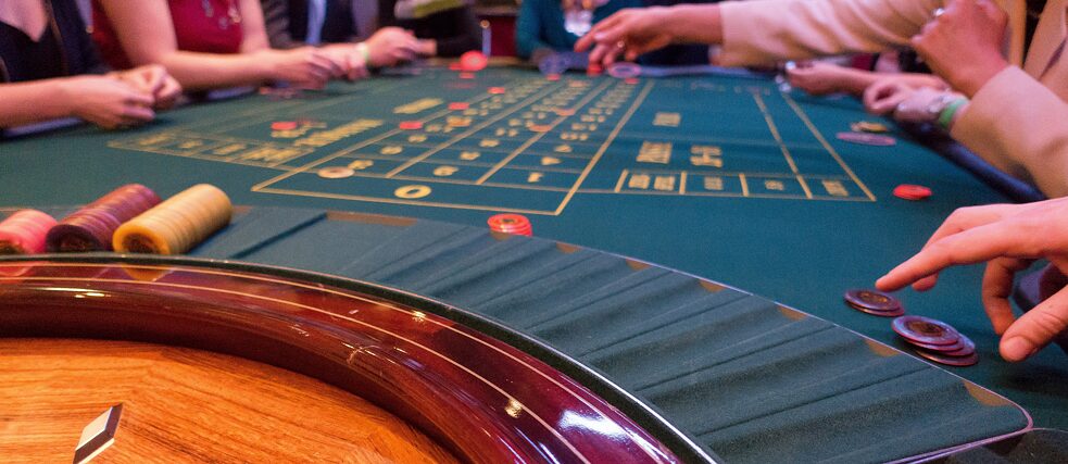 Roulettetisch mit einem langen, mit grünem Tuch (Tapis) bespannt, Jetons und Hände im Hintergrund.