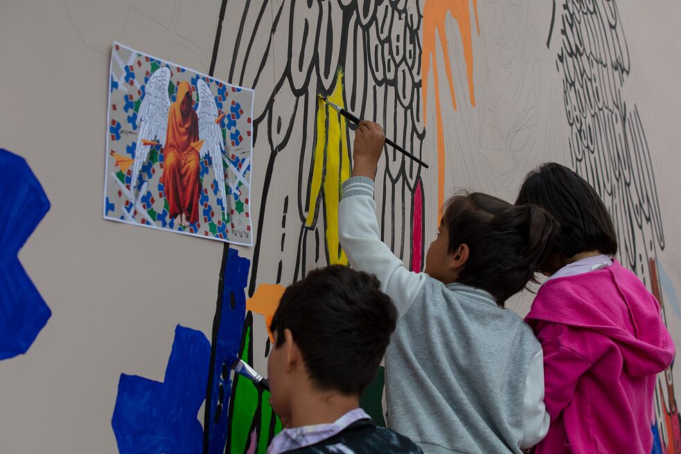An einer großen Wand malen drei Kinder Farbflächen aus. Links neben ihnen hängt die Skizze des fertigen Kunstwerks.