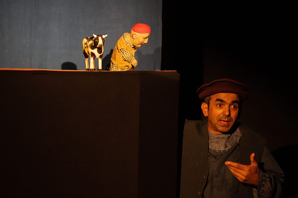 Eine Kuh und eine Puppenfigur stehen auf einer erhöhten Bühne. Rechts vor steht der Puppenspieler Abdul Haq Haqjoo.