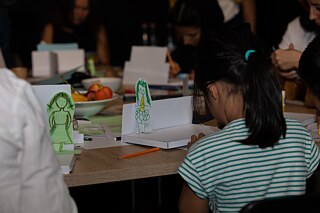 A child is shown sitting at the table from behind. On the table are self-made stages and figures, pens and paper.