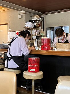 Two people wearing breathing masks sit opposite each other at a counter, both seem to be very concentrated on one task each.