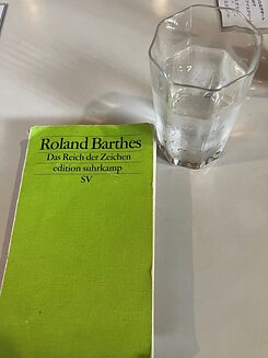 Closed book with the inscription “Roland Barthes - In the Realm of Signs”, next to it a glass with a clear liquid