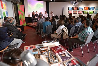 The participants of the panel discussion can be seen in the background. The audience is sitting in front of them. In the foreground of the picture, a bookstall can be seen out of focus.