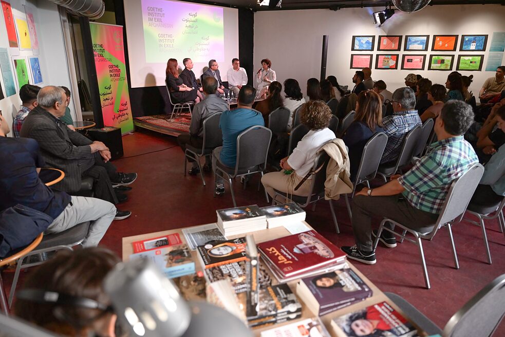 The participants of the panel discussion can be seen in the background. The audience is sitting in front of them. In the foreground of the picture, a bookstall can be seen out of focus.