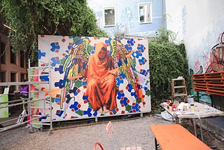 Ein großes Wandgemälde ist zu erkennen. Darauf sieht man ein Mädchen in orangefarbenem Gewand. Sie trägt bunte Flügel. Im Hintergrund ist eine grün-blau rote Ornamentik zu erkennen. Rechts neben dem Bild steht ein Tisch mit Farbtuben. Links davon steht ein Rollgerüst.