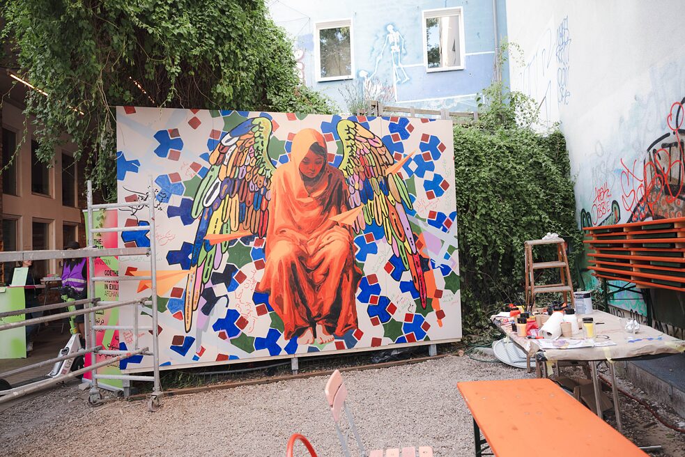 Ein großes Wandgemälde ist zu erkennen. Darauf sieht man ein Mädchen in orangefarbenem Gewand. Sie trägt bunte Flügel. Im Hintergrund ist eine grün-blau rote Ornamentik zu erkennen. Rechts neben dem Bild steht ein Tisch mit Farbtuben. Links davon steht ein Rollgerüst.