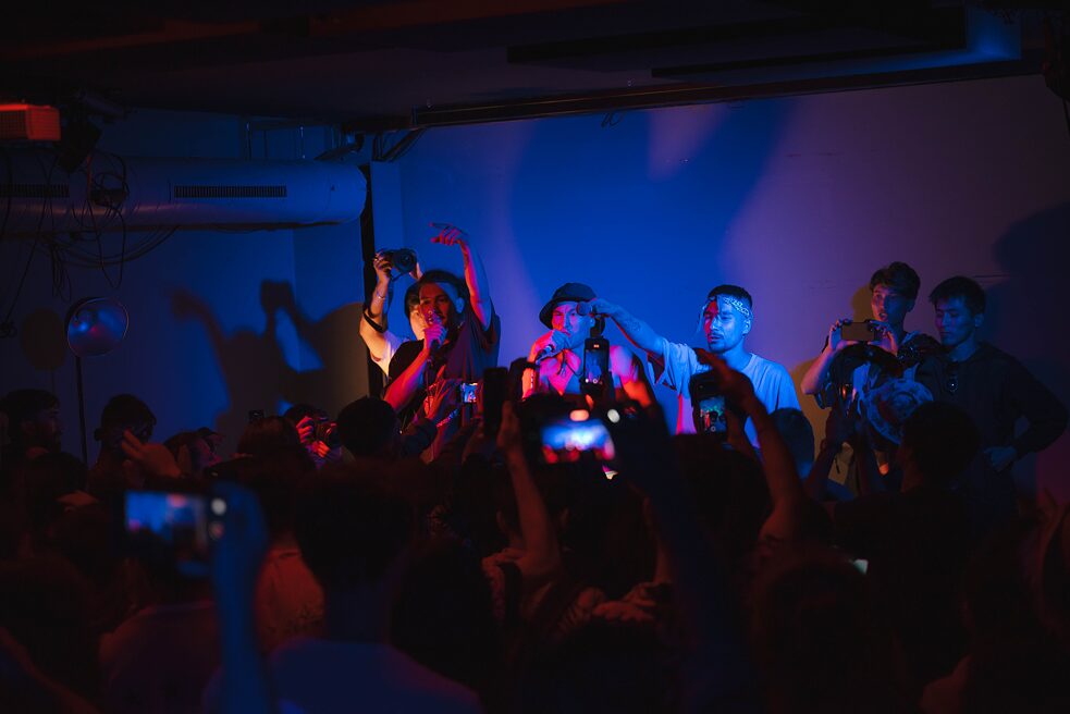 3 Mitglieder von AK13 sind auf der Bühne zu sehen. Während die zwei linken Männer rappend dargestellt sind, hält der rechte Mann sein Mikrofon in Richtung Publikum.