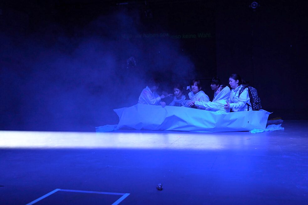 A group of young people sit in a boat made of paper. The room is lit in blue and fog rises.