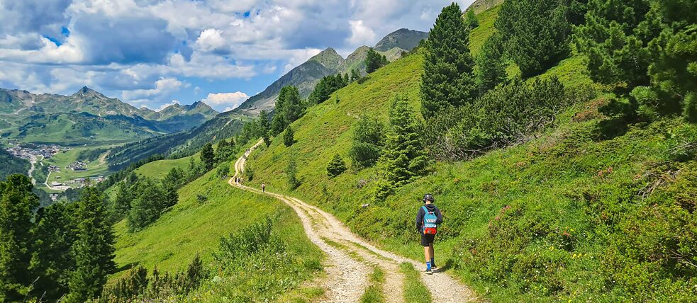 Jugendlicher auf einem Wanderweg in den Bergen. Er trägt große Kopfhörer und einen kleinen Rucksack.