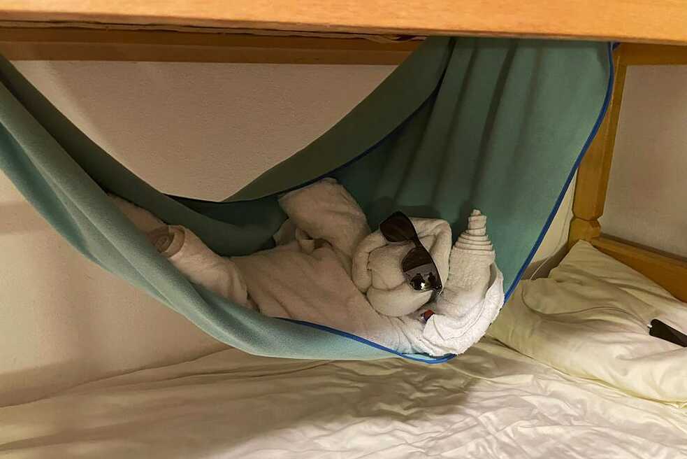 In einem Stockbett liegt in einer auf einer Hängematte (Strandtuch) eine aus weißen Handtücher kreierte Figur. Sie trägt eine Sonnenbrille.
