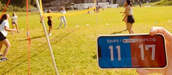 Im Vordergrund ein Handy mit einer Spielanzeige auf Spanisch 11 zu 17. Im Hintergrund Jugendliche beim Volleyball