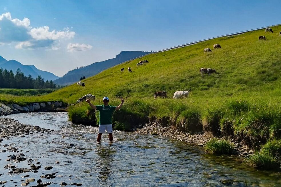 Ein junger Mann steht in einem Bach bis zu den Waden im Wasser und hält die Arme nach oben.