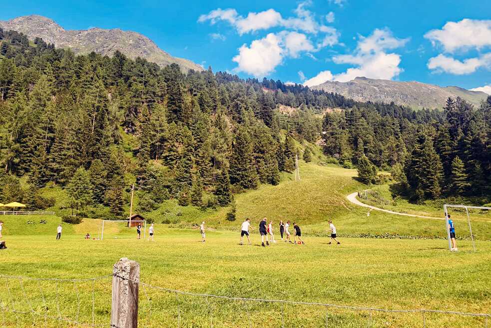 Jugendliche spielen Ball auf einer großen Wiese. Im Hintergrund sind Berge und Wälder zu sehen.