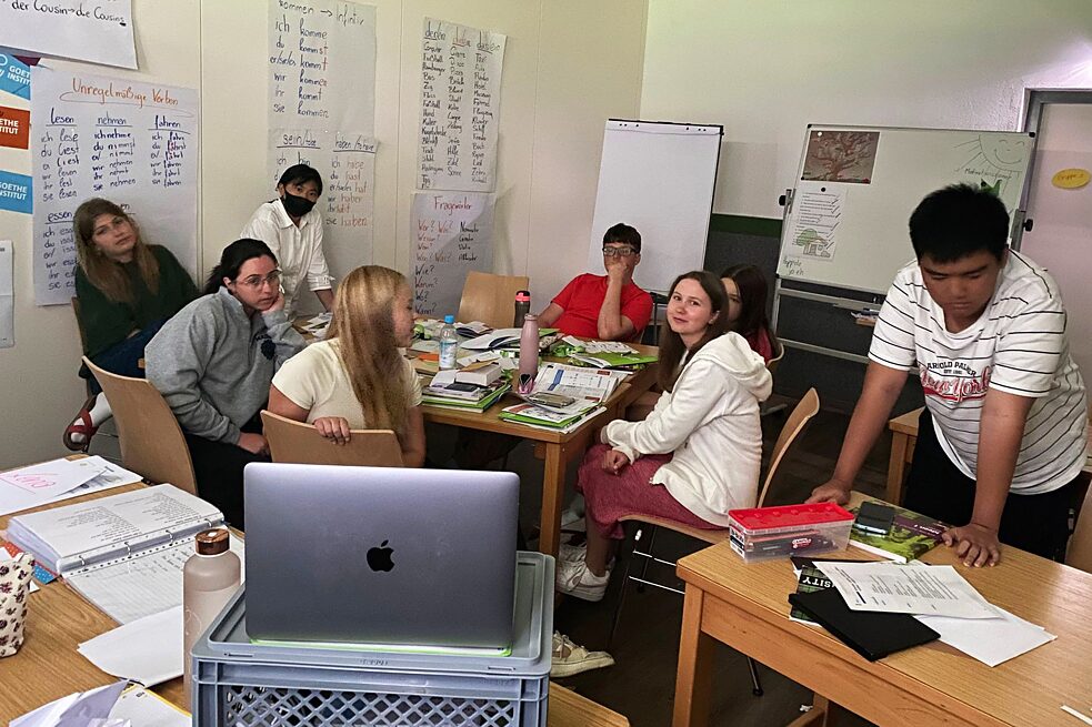 Jugendliche in einem Klassenraum. An den Wänden hängen Plakate, im Vordergrund steht ein aufgeschlagener Laptop.