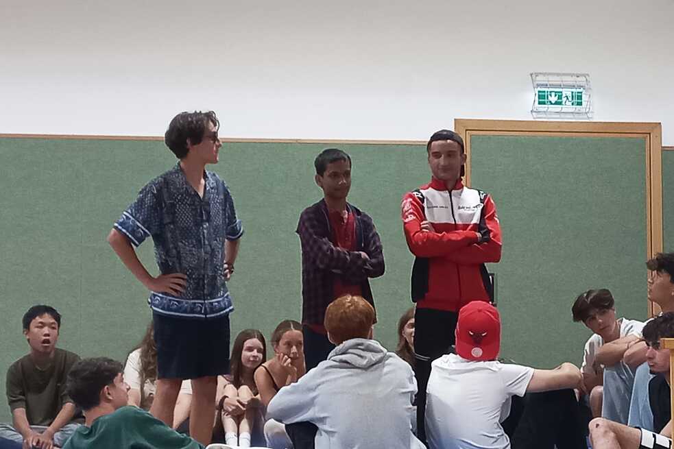 Drei Jugendliche stehen mitten in einer Gruppe auf dem Fußboden einer Sporthallte sitzender Jugendlicher.