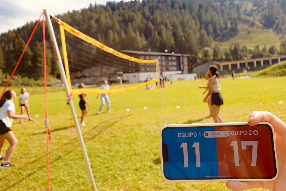 Im Vordergrund ein Handy mit einer Spielanzeige auf Spanisch 11 zu 17. Im Hintergrund Jugendliche beim Volleyball