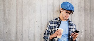 Ένας νεαρός άνδρας με μπλε μπερέ και καρό πουκάμισο κρατά στο χέρι του ένα smartphone και φορά ακουστικά in-ear.