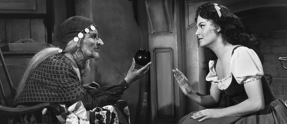 Ein schwarz-weißes Filmstill, rechts sieht man die Hexe, welche Schneewitchen, links sitzend, einen Apfel reicht.