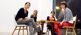 Vier junge Frauen sitzen an einem Tisch und lächeln in die Kamera.