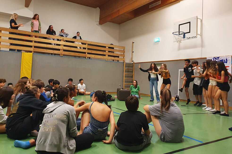 Jugendliche in einer Turnhalle sitzen auf dem Fußboden, andere stehen und singen.