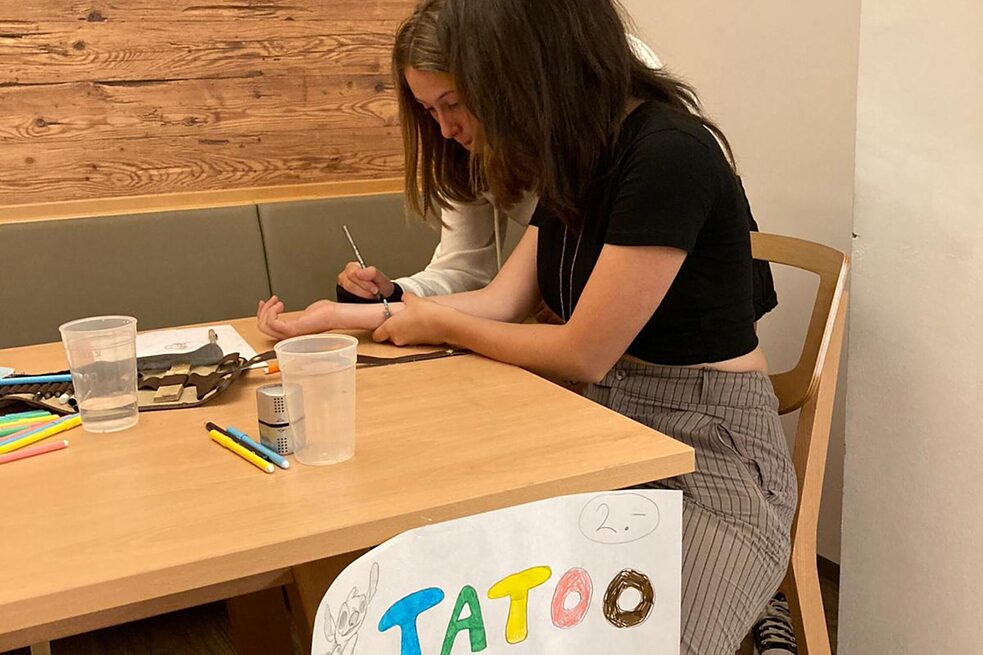 Jugendliche an einem Tisch tätowieren einen Unterarm.