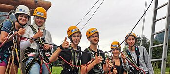 Jugendliche mit Helm und gut gesichert im Kletterpark lächeln in die Kamera und zeigen Daumen hoch.
