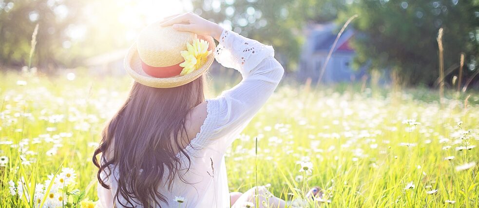 Jugendliche mit Strohut und Blume sitzt auf einer Wiese im Gras.