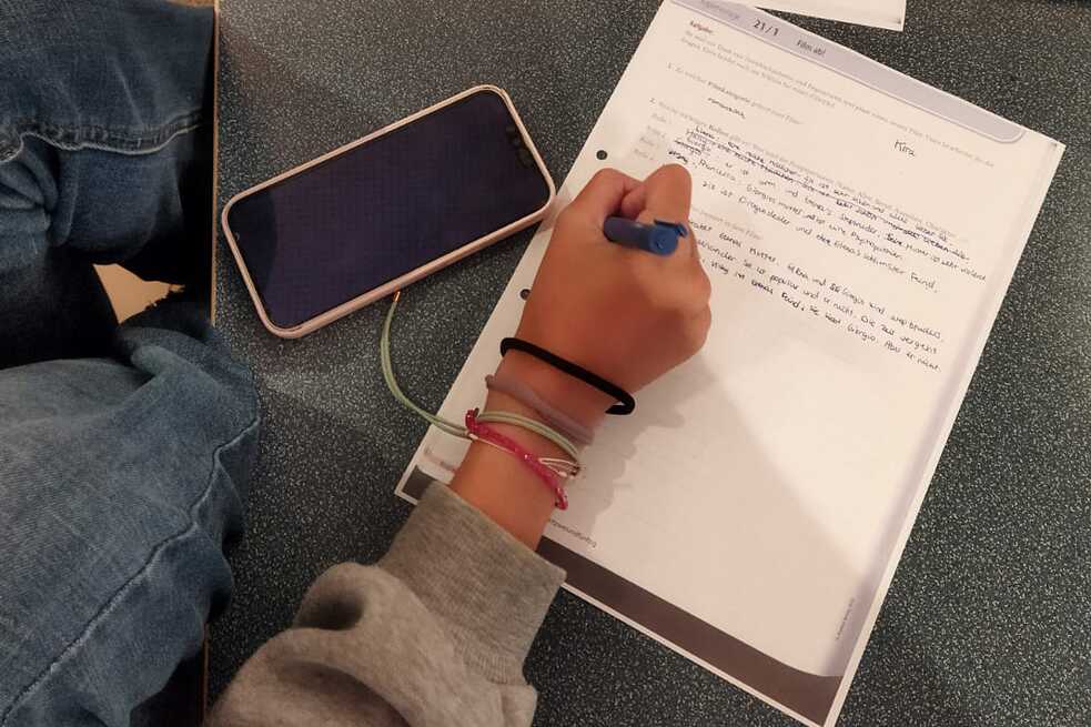 Ein Arbeitsblatt wird von einer Jugendlichen ausgefüllt. Ein Smartphone liegt neben der Vorlage auf dem Tisch.