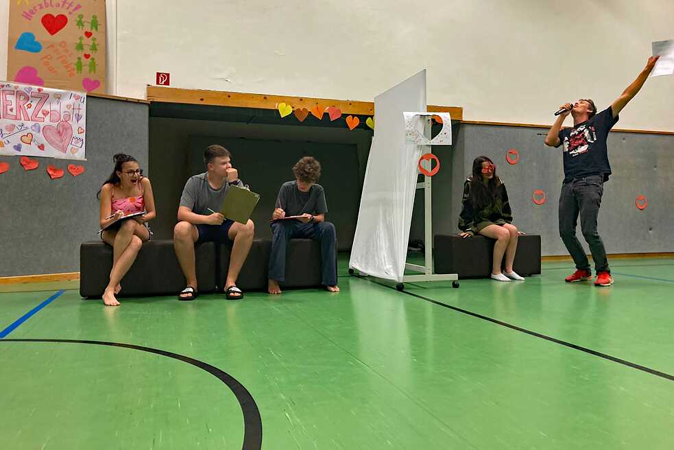 Jugendliche in einer Turnhalle spielen die Sendung „Herzblatt“ nach.