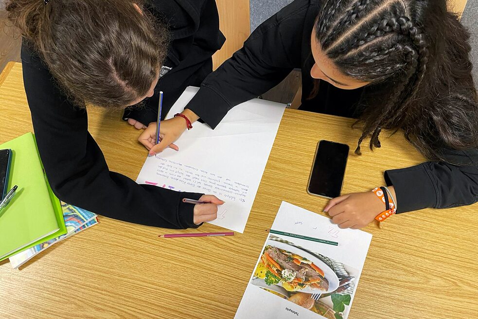 Zwei Jugendliche an einem Tisch bearbeiten ein Rezept.