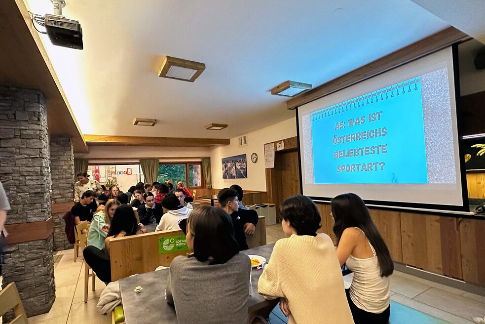 Jugendliche an Tischen, ein Projektor wirft eine Quizfrage auf eine Leinwand.