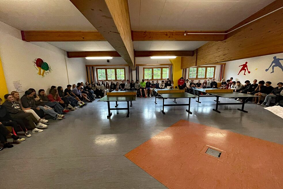 Jugendliche sitzen im Kreis in einem sehr großen Raum.