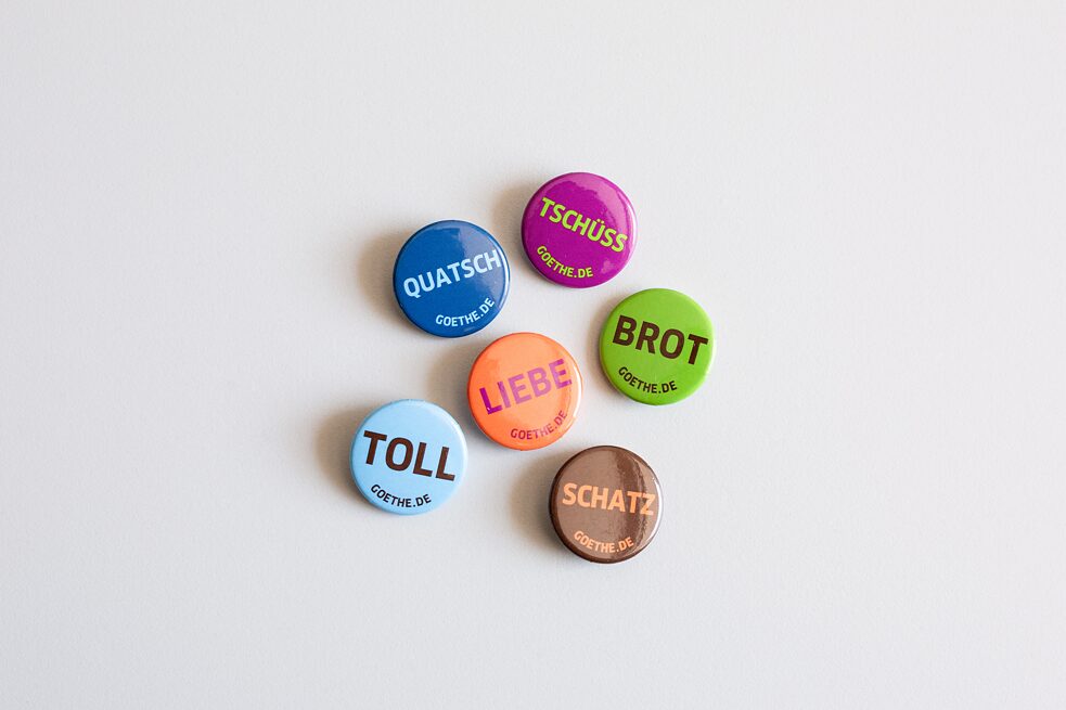 6 badges in light blue, dark blue, purple, brown, orange and green with the German words Quatsch, toll, Liebe, tschüss, Btor and Schatz on it. 