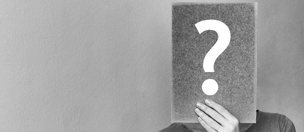 Jemand hält sich ein Karton mit einem Fragezeichen vor das Gesicht.