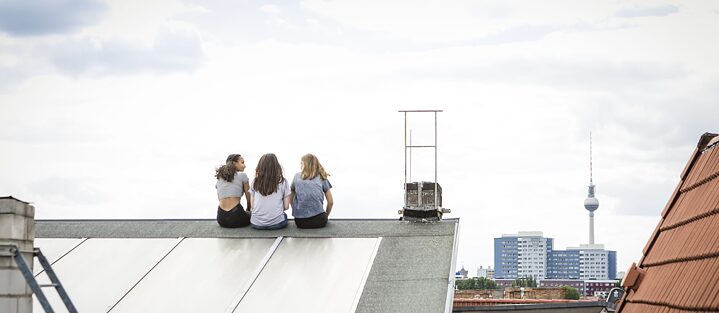 Rückansicht auf drei Freundinnen, die auf einem Dach in Berlin sitzen und sich unterhalten. Im Hintergrund der Fernsehturm.