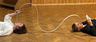 Zwei Performer*innen liegen auf dem Rücken auf dem Boden, haben einen Flexiply-Streifen im Mund und ihr Handy in der Hand