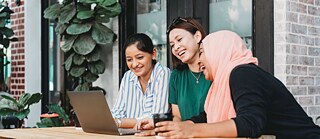 Три девушки сидят на улице за столом. Перед ними ноутбук. Скорее всего у них видеоконференция.