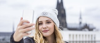 Eine junge Frau mit grauer Mütze fotografiert sich lächelnd mit dem Smartphone. Im Hintergrund der Kölner Dom.