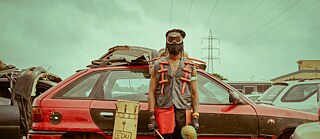 Ein Mann in einer seltsamen Aufmachung vor einem Autowrack. Der Mann hält in seiner linken Hand ein traditionelles Musikinstrument.