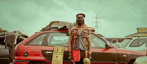 Ein Mann in einer seltsamen Aufmachung vor einem Autowrack. Der Mann hält in seiner linken Hand ein traditionelles Musikinstrument.