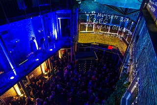 Der mit Menschen gefüllte Innenhof ist von oben fotografiert und zeigt die Video-Installation an den Wänden. Dort steht "Wir müssen diesen Krieg gemeinsam gewinnen".