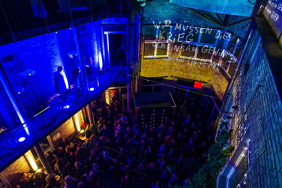 Der mit Menschen gefüllte Innenhof ist von oben fotografiert und zeigt die Video-Installation an den Wänden. Dort steht "Wir müssen diesen Krieg gemeinsam gewinnen".