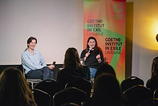 Schriftsteller Roman Malynowsky sitzt links lächelnd neben der Moderatorin Evgenia Lopata, die gerade ins Mikrofon spricht und dabei in das Publikum blickt. 