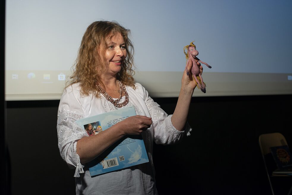 Lana Ra hält ihr Buch im rechten Arm. Mit der linken Hand hält sie ein Pferdefigürchen hoch