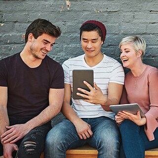 Eine Gruppe junger Leute sitzen nebeneinander auf eine Bank. Sie lesen von Laptops und Tablets.